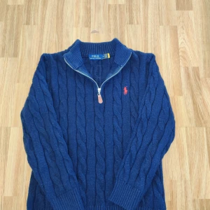 Mörkblå stickad tröja med dragkedja från Polo Ralph Lauren - Mörkblå kabelstickad tröja från Polo Ralph Lauren med halv dragkedja och röd broderad logga på bröstet. Tröjan har hög krage och ribbade muddar vid ärmslut och nederkant. Perfekt för en stilren och klassisk look.