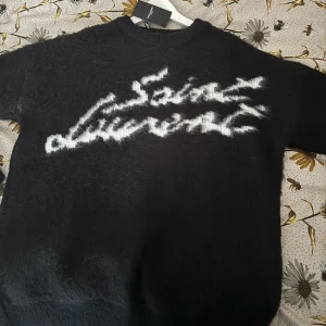 Svart stickad tröja från Saint Laurent - Säljer en svart stickad tröja från Saint Laurent med vit, suddig logotyp framtill. Tröjan har långa och en rund halsringning. Perfekt för dig som gillar exklusiva och trendiga plagg med statement-tryck. Tröjan är dessutom aldrig använd och säljs för att den inte passar.