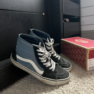 Vans Sk8-Hi sneakers i svart och blått - Säljer ett par klassiska Vans Sk8-Hi sneakers i svart och ljusblå mocka med vit sula och vit sidosnirk. Höga skaft och vita snören. 