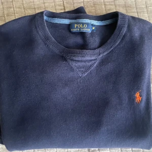 Mörkblå crewneck från Polo Ralph Lauren - Säljer en klassisk mörkblå tröja från Polo Ralph Lauren med rund halsringning och den ikoniska broderade loggan i orange på bröstet. Tröjan har långa ärmar och är tillverkad i mjukt material, perfekt för en stilren vardagslook. Stlk M. 9/10 skick knappt andvänd då jag snabbt växte ur storleken.