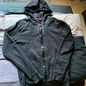 Svart hoodie och byxa med dragkedja från Hugo - Säljer en svart hoodie och byxa från Hugo med dragkedja framtill och huva med snörning. Tröjan har ribbade muddar vid ärmslut och nederkant samt snygga detaljer med svarta ränder längs dragkedjan. Perfekt för en avslappnad stil.