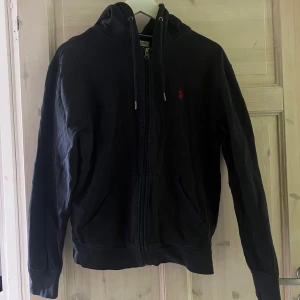 Svart hoodie med dragkedja från U.S. Polo Assn. - Svart hoodie från U.S. Polo Assn. i storlek M. Tröjan har dragkedja framtill, huva med snören och en liten röd broderad logga på bröstet. Perfekt för en avslappnad stil.