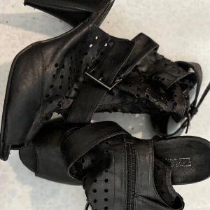 Svarta sandaler med klack och perforerat mönster - Svarta sandaler från Exani med klack, perforerat mönster och dragkedja på sidan. Skorna har öppen tå och justerbart spänne vid vristen. Perfekta för dig som vill ha en stilren och luftig sko med snygga detaljer.