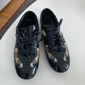 Burberry sneakers med klassiskt rutmönster - Säljer ett par snygga Burberry sneakers med det ikoniska beige rutmönstret och svarta lackdetaljer. Skorna har snörning och rund tå, perfekt för dig som vill ha en stilren och tidlös look.