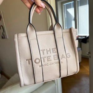 Beige handväska från Marc Jacobs - Säljer en beige handväska från Marc Jacobs med texten 'THE TOTE BAG' på framsidan. Väskan har två handtag och är tillverkad i ett strukturerat syntetmaterial. Perfekt för att bära det viktigaste i vardagen.