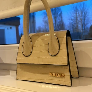 Beige handväska  - Superstylish beige handväska från med croco-mönstrad yta och guldigt logodetalj framtill. Väskan har ett handtag. Band medföljer. Helt oanvänd ✨