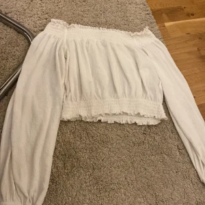 Vit offshoulder topp från H&M Divided - Säljer en söt vit offshoulder topp från H&M Divided i storlek XS. Toppen har långa ärmar, smockdetaljer upptill och nertill samt en lätt volangkant längst ner. Perfekt för en trendig och luftig look!