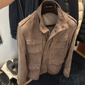 Rose & Born mockajacka - Snygg beige mockajacka med fyra framfickor och axelklaffar. Jackan har hög krage och stängs med både dragkedja och knappar. Perfekt för dig som vill ha en stilren och tidlös look. Kvitto följer med 