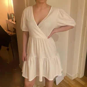 Vit omlottklänning med puffärm - Säljer en söt vit kortklänning med omlottliv och djup v-ringning. Klänningen har puffiga kortärmar och volang i kjolen för en luftig känsla. Perfekt för sommarens alla tillfällen! Perfekta studentklänningen❤️