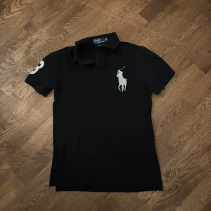 Svart pikétröja från Polo Ralph Lauren - Svart pikétröja från Polo Ralph Lauren med vit broderad logga på bröstet och siffra på ärmen. Slim fit-modell med klassisk krage och korta ärmar. Perfekt för en stilren och sportig look. Säljer för att det är inte min smak. Fick av brorsan. Det är min storlek så det är bara att skriva om du har några funderingar 😁Pris kan diskuteras!