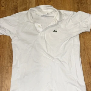 Vit piké från Lacoste - Säljer en klassisk vit pikétröja från Lacoste med broderad grön krokodil på bröstet. Tröjan har korta ärmar, krage och knappar framtill. Perfekt för en stilren och sportig look.