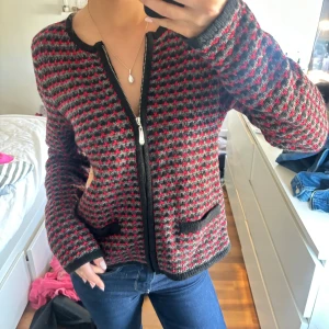 Mönstrad stickad jacka från Isolde - Säljer en stickad jacka från Isolde i svart, grått och rött med dragkedja framtill och två fickor. Jackan har lång ärm och ett diskret mönster som ger en cool retrovibe. Perfekt för dig som gillar unika plagg med lite edge.