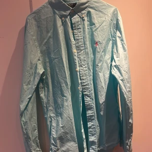 Ljusblå skjorta från Polo Ralph Lauren - Säljer en ljusblå långärmad skjorta från Polo Ralph Lauren i slim fit-modell. Skjortan har klassisk krage, vita knappar och en broderad rosa logga på bröstet. Perfekt för dig som gillar stilrena och klassiska plagg.