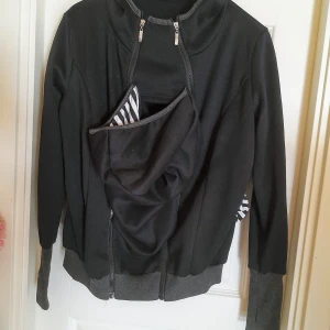 Svart hoodie med bärinsats och zebramönster - Svart hoodie med grå muddar och huva. Har en smart bärinsats med svartvitt zebramönstrat foder, perfekt för att bära barn. Dragkedjor framtill för enkel öppning och stängning. Mjuk och bekväm, passar till vardags. Jag tror att den är hemsydd då det inte finns lappar i. Bärtröja. Bärjacka. Den passar en small. Eventuellt en medium med. Använd en gång 