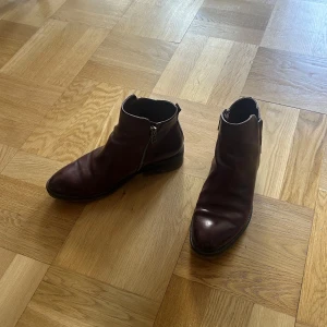 Vinröda 90-tals boots med dragkedja - Säljer ett par vinröda boots i skinn med rund tå och dragkedja på sidan. Skorna har en klassisk design och låg klack, perfekta för dig som gillar stilrena och tidlösa skor.