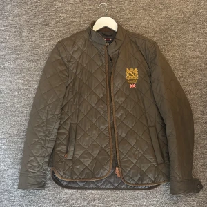 Olivgrön quiltad jacka från Morris - Snygg olivgrön quiltad jacka från Morris med ståkrage och broderad logga på bröstet. Jackan har bruna detaljer, fickor framtill och stängs med dragkedja. Perfekt för dig som gillar klassisk stil med modern touch.