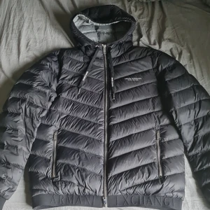Svart pufferjacka från Armani Exchange - Säljer en svart pufferjacka från Armani Exchange med huva och dragkedja. Jackan har diskret logga på bröstet och bred resår med logotyp nertill. Perfekt för kyliga dagar och riktigt snygg streetstil. Skick är 10/10 finns inga defekter. Ny pris ligger runt 2800kr mitt pris 1379 som sagt den är 10/10 i skick
