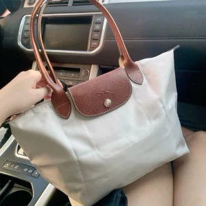  handväska från Longchamp med bruna detaljer - Säljer en klassisk vit handväska från Longchamp med bruna handtag och lock i skinn. Väskan har guldknapp och är perfekt för att bära dina vardagsprylar. Enkel och stilren design som passar till många outfits.