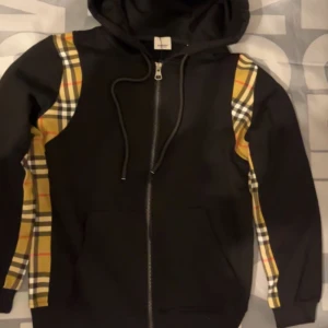 Svart hoodie med rutig detalj - Svart hoodie med dragkedja och huva. Snygga rutiga paneler i beige, rött och gult längs axlar och ärmar. Perfekt för en trendig streetwear-look. Dragsko i huvan och ribbade muddar.