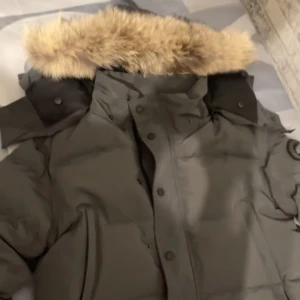 Svart dunjacka med pälskrage - Säljer en svart dunjacka med stor, fluffig pälskrage och hög krage. Jackan har knappar och dragkedja framtill samt en patch på ärmen. Perfekt för kalla dagar.