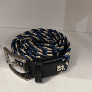 Flätat blått och beige bälte från BM - Snyggt flätat bälte i blått, beige och mörkblått med silverfärgat spänne. Bältet har detaljer i svart skinn och en metallbricka med BM-logga vid spännet. Perfekt accessoar för att lyfta din outfit.