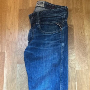 Blå jeansbyxor från Replay - Säljer ett par klassiska blå jeans från Replay med snygga slitningar och femficksdesign. Jeansen har normal passform och tydliga detaljer vid fickorna. Perfekta för en avslappnad stil.