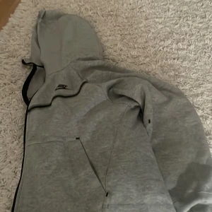 Grå hoodiejacka från Nike - Grå hoodiejacka från Nike med svart dragkedja och svarta detaljer. Jackan har huva och två fickor framtill. Perfekt för en sportig och avslappnad stil. Aldrig använd säljer för den e för stor nypris 1100kr