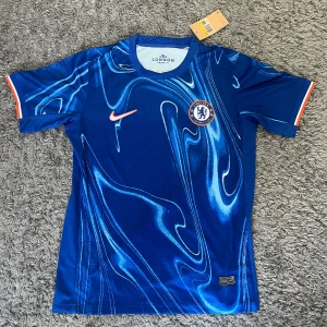 blå Chelsea tröja från Nike - Storlek M. blå Chelsea fotbollströja från Nike med mönster i olika blå nyanser och orange detaljer. Tröjan har klubbmärke på bröstet och Nike logga. Nästan helt oanvänd och helt ren. Inget namn på ryggen. PRISET KAN DISKUTERAS.