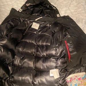 Svart dunjacka från Moncler - Säljer en svart dunjacka från Moncler med huva och broderad Moncler-logga på ärmen. Jackan har dragkedja framtill och ett unikt serietidningsmärke på insidan. Klassisk pufferstil med quiltad design och röd detalj vid dragkedjan. OBS dragkedjan är trasig lätt att fixa där av priset ! 