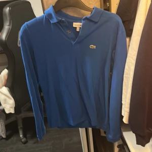 Blå långärmad pikétröja från Lacoste - Säljer en klassisk blå långärmad pikétröja från Lacoste med den ikoniska krokodilloggan på bröstet. Tröjan har krage och knäppning vid halsen, perfekt för en stilren och sportig look.