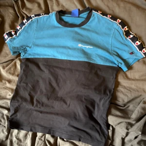 Champion t-shirt med blå och svart färgblock - Snygg t-shirt från Champion med blått upptill och svart nedtill. Logga broderad på bröstet och coola band med Champion-loggor längs axlarna. Rund halsringning och korta ärmar. Perfekt för en sportig look.