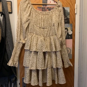 Blommig volangklänning med lång ärm - Supersöt kortklänning med småblommigt mönster i beige och brunt. Klänningen har långa ärmar, volangdetaljer och en smickrande v-ringning. Perfekt för vår och sommar, med en luftig och romantisk känsla.