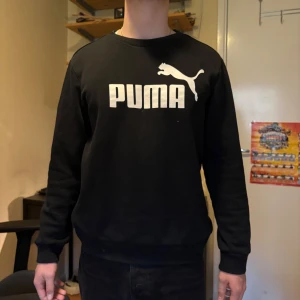 Svart sweatshirt från Puma - Säljer en svart sweatshirt från Puma med stor vit logga framtill. Tröjan har rund halsringning och långa ärmar med ribbade muddar. Perfekt för en avslappnad stil.