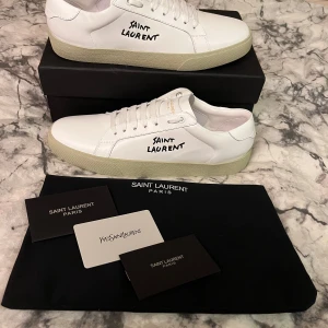 Vita Saint Laurent sneakers med svart text - Säljer ett par stilrena vita sneakers från Saint Laurent med svart handskriven logga på sidan. Skorna har rund tå, platt sula och är tillverkade i skinn. Perfekta för dig som gillar minimalistisk och lyxig design.