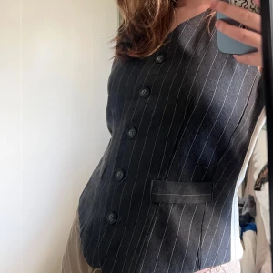 Randig väst från Necessary Objects - Snygg mörkgrå väst med vita ränder från Necessary Objects. Klassisk knäppning framtill med fem knappar och två fickor. Västen har en justerbar rem i ryggen. Perfekt för både stilren eller edgy look.
