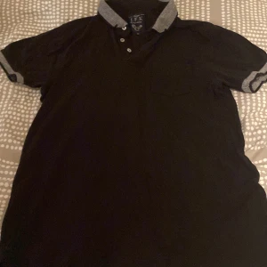 Svart pikétröja med grå krage från J.F.K - Säljer en svart pikétröja från J.F.K med grå detaljer på krage och ärmslut. Tröjan har korta ärmar, knappar vid halsen och en liten bröstficka. Perfekt för en stilren och avslappnad look.