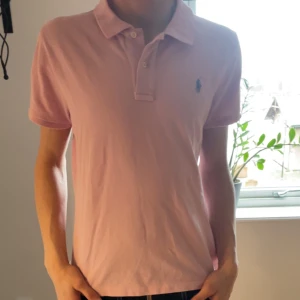 Polo tröja - Säljer en stilren rosa t-shirt från Ralph Lauren med korta ärmar och klassisk krage. Den har en broderad logga i blått på bröstet och knappar vid halsen. Perfekt för en avslappnad look!