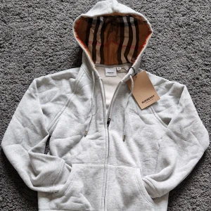Burberry zip hoodie med klassiskt rutigt foder - Säljer en grå hoodie från Burberry med dragkedja och fickor framtill. Insidan av huvan har det ikoniska Burberry-rutiga fodret. Broderad logga på bröstet och snygga detaljer vid snörningen. Perfekt för en stilren och avslappnad look. Storlek M men passar S/XS (passar nån på 165-173 cm skulle jag säga) Nypris 8000 kr Mitt pris 1000 kr (går att sänka någon hundring). Skriv DM vid fråga! 🤩💯
