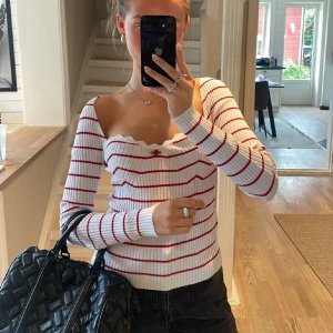 Vit ribbad tröja med röda ränder och volangkant - Säljer en jättesöt vit tröja med röda ränder. Tröjan har en söt volangkant vid halsringningen och en liten röd ros framtill. 💕