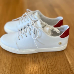 Vita sneakers med röd detalj från Lauren Ralph Lauren - Snygga vita sneakers i skinn från Lauren Ralph Lauren med röd detalj på hälen och guldfärgad logga på sidan. Klassisk modell med snörning och rund tå. Perfekta för en stilren och sportig look. Helt oanvända! Så nyskick