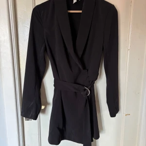 Svart byxdress med bälte - Säljer en svart byxdress med långa ärmar och djup v-ringning. Ser ut som en kort klänning framifrån och shorts bakifrån