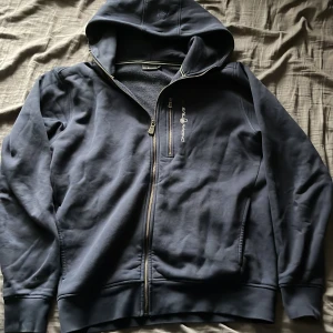 Mörkblå hoodie från Sail Racing - Säljer en mörkblå hoodie från Sail Racing med dragkedja framtill och en extra bröstficka med dragkedja. Tröjan har huva och broderad logga på bröstet. Perfekt för dig som gillar sportig och stilren design. Mycket bra skick 9/10