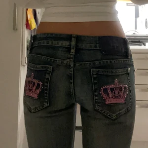 Victoria Beckham jeans - Säljer ett par mörkblå jeans som är bootcut och snygga rosa kron-motiv på bakfickorna. Jeansen har låg midja och klassisk femficksdesign. Perfekta för dig som vill sticka ut med coola detaljer. Säljer då de är precis förkorta på mig som är 171cm🩷❤️
