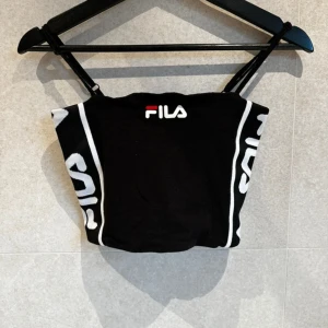 Svart tubtopp från FILA - Svart tubtopp från FILA med vita detaljer och logga framtill. Toppens sidor har vita ränder och texten 'FILA' i vitt. Perfekt för en sportig och trendig look.