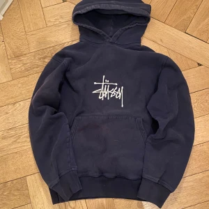 Stüssy hoodie - Säljer en mörkblå hoodie från Stüssy med vit logga broderad på bröstet. Tröjan har huva, magficka och ribbade muddar på ärmar och nederkant. Perfekt för en avslappnad streetwear-look. Lite solblekt på framsidan (se bild 3) men annars inga defekter. Storlek medium.  Nypris 1600 kr.                                                                                 Skriv gärna vid frågor eller funderingar!