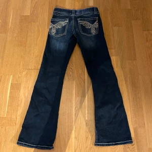 Mörkblå jeansbyxor med broderade bakfickor från Gina Tricot - Snygga mörkblå jeansbyxor från Gina Tricot med kontrastsömmar och broderade detaljer på bakfickorna. Byxorna har bootcut-modell och klassisk femficksdesign. Perfekta för dig som gillar en cool och tidlös stil.