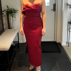 Vinröd långklänning med draperad överdel - Säljer en elegant vinröd långklänning med draperad, avlappnad överdel och smal passform. Klänningen har slits på baksidan och är ärmlös, perfekt för festliga tillfällen. Materialet känns mjukt och följsamt.