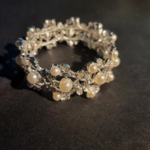Silverfärgat armband med pärlor och strass - Säljer ett glittrigt armband i silverfärg med vita pärlor och gnistrande stenar. Armbandet har ett romantiskt och detaljerat mönster som ger en lyxig känsla. Perfekt för dig som vill sticka ut med ett unikt smycke.
