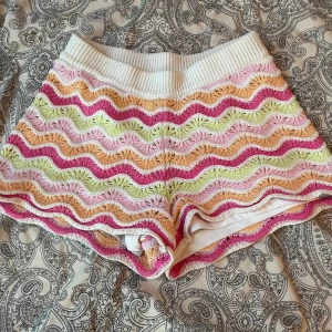 Färgglada virkade shorts med vågmönster - Superfina virkade shorts med vågigt mönster i rosa, orange, grön och vitt. De har en bred, ribbad midja och är perfekta för sommaren. Mjukt material och loose fit gör dem både bekväma och trendiga. Storlek 134-140 i barnstorlek 
