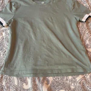 Grön t-shirt från H&M med kontrastdetaljer - Snygg grön t-shirt från H&M med vita och svarta kontrastdetaljer på ärmsluten. Klassisk rund halsringning och rak passform. Perfekt för en avslappnad stil. Storlek 158-164 i barnstorlek